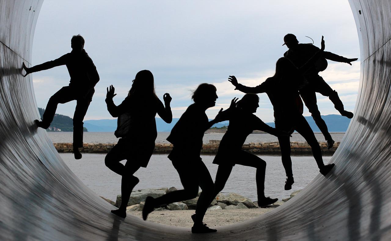 Silhouettes of a group of 6 friends running up the walls of what appears to be a giant pipe. Mountains and a blue sky in the background.